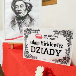 Na zdjeciu widac dekorację na "Narodowe Czytanie" w DPS. Na przodzie widac plakat informacyjny, a w tle widac podobiznę Adama Mickiewicza i biało-czerwoną flagę